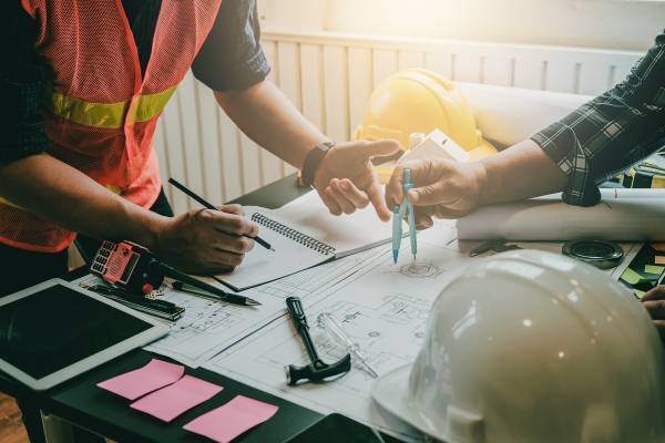 Construction professionals reviewing blueprints and plans on desk with safety vest, hard hats, and tablet device during project planning meeting