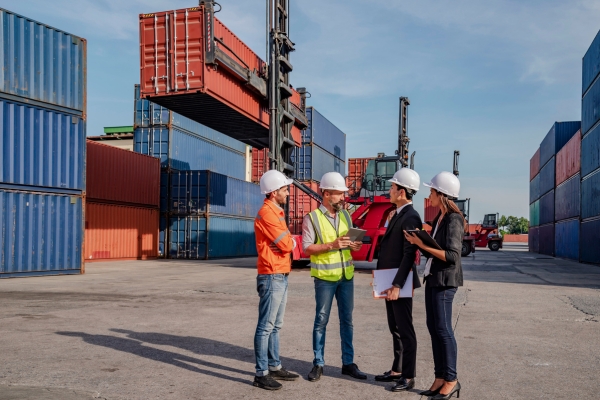 Team of logistics professionals discussing operations in container terminal yard, wearing safety equipment and reviewing documents