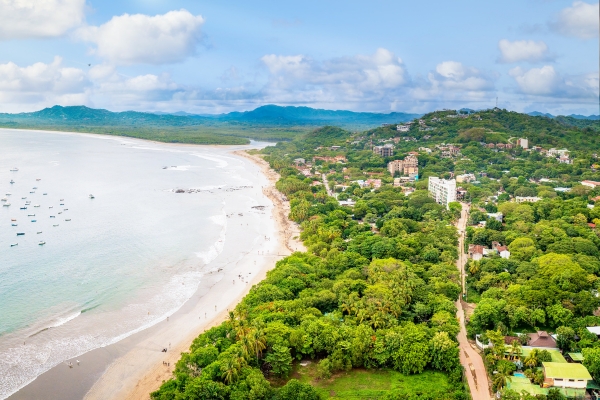 Aerial drone photograph of Tamarindo Beach showing curved coastline, lush green vegetation, beachfront properties, and calm Pacific waters with boats