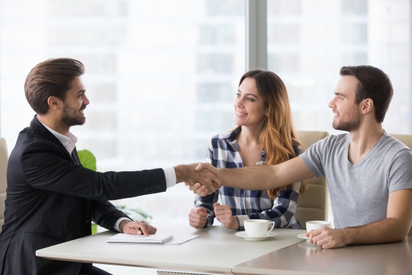 Professional in suit shaking hands with casual-dressed client couple during business meeting at bright office table with coffee