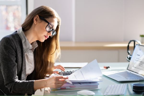 Woman in glasses reviewing financial paperwork, calculator, laptop at workspace