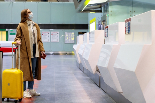 Woman wearing face mask standing at empty airport check-in counters with yellow luggage, wearing brown coat and following safety travel protocols