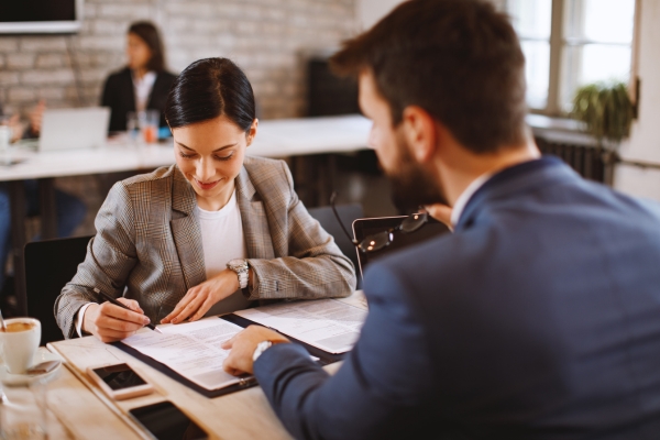 Two business professionals reviewing documents at a meeting table, woman in gray blazer and man in navy suit discussing paperwork over coffee